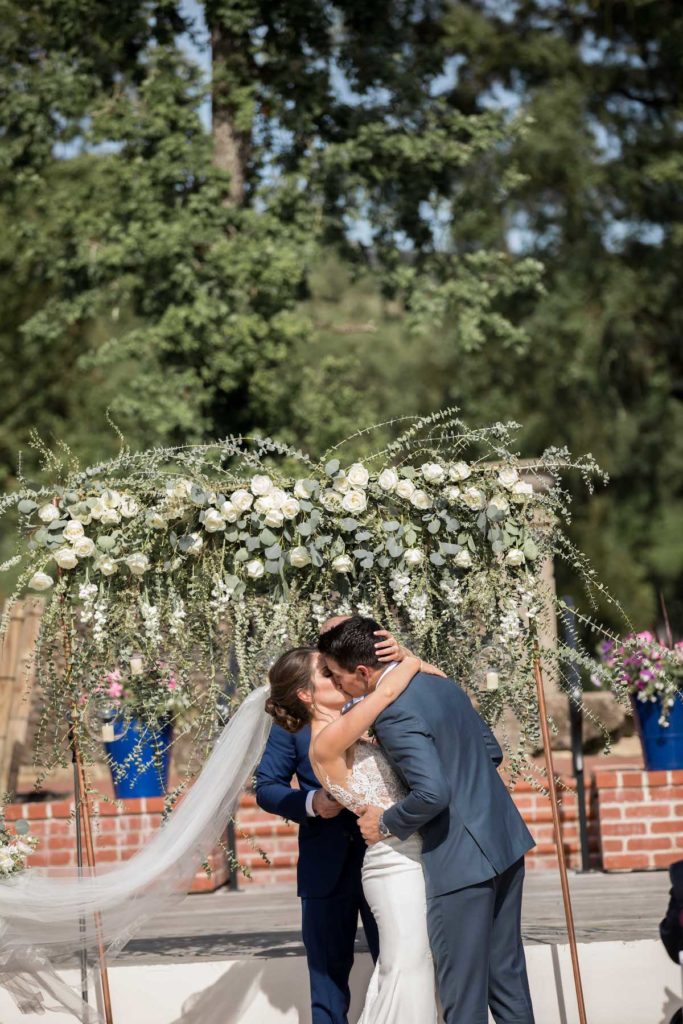 Bride and groom having first kiss in front of green & white floral arch